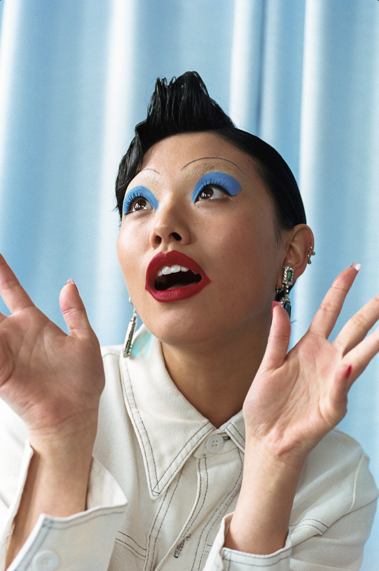 Eye-level portrait of a woman with blue eyeshadow, red lipstick, and a white shirt against a light blue background, looking upwards.