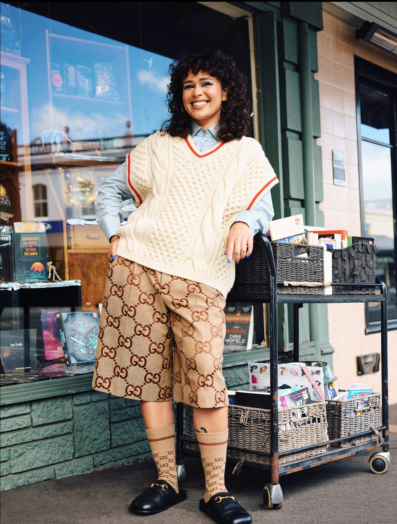 A smiling adult woman poses outdoors wearing designer shorts, socks, sweater vest, and slides, standing beside a cart of books and goods.