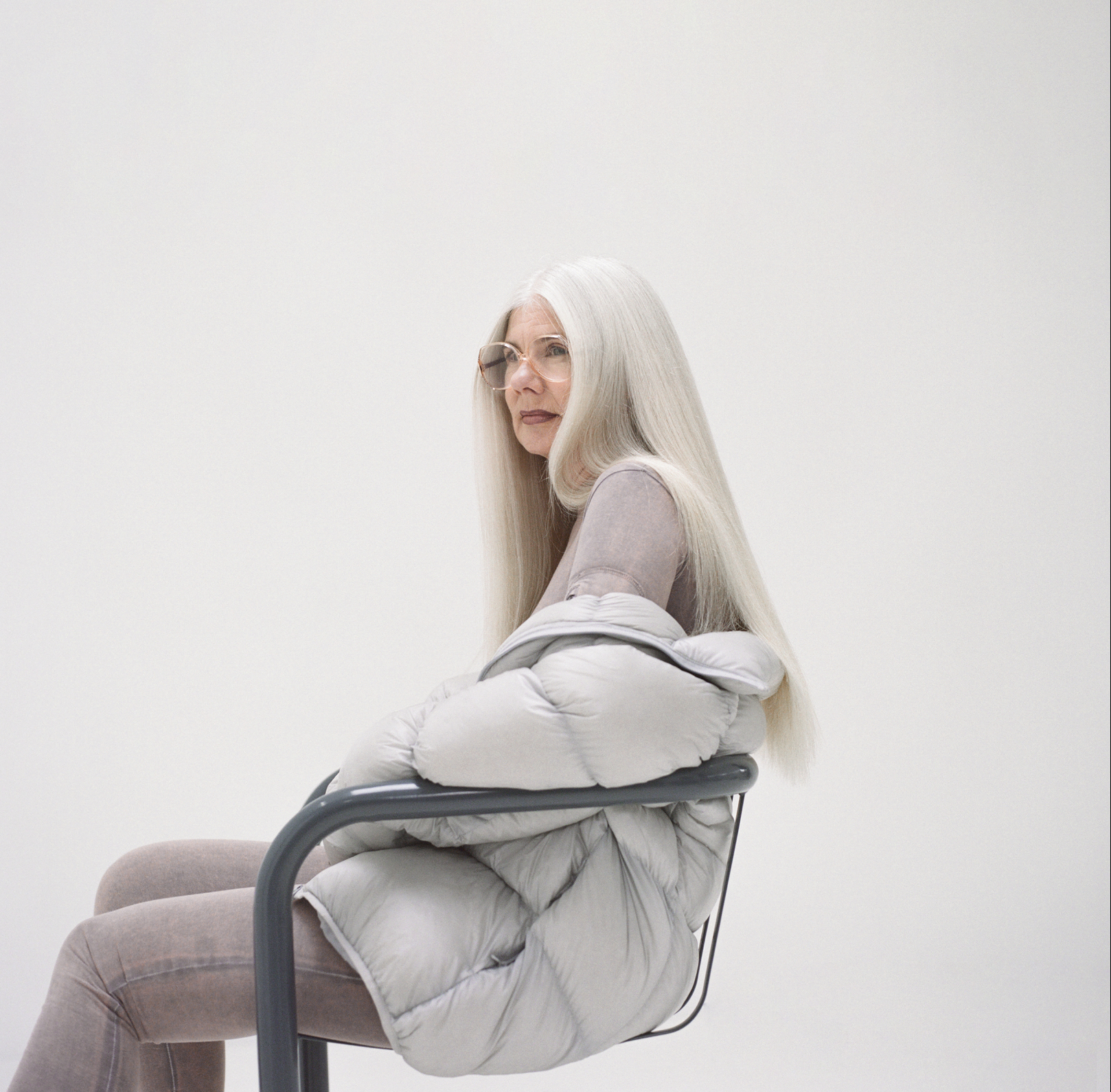 Elegant mature woman with long gray hair, wearing glasses and a padded jacket, sits in a gray chair against a clean, bright background.