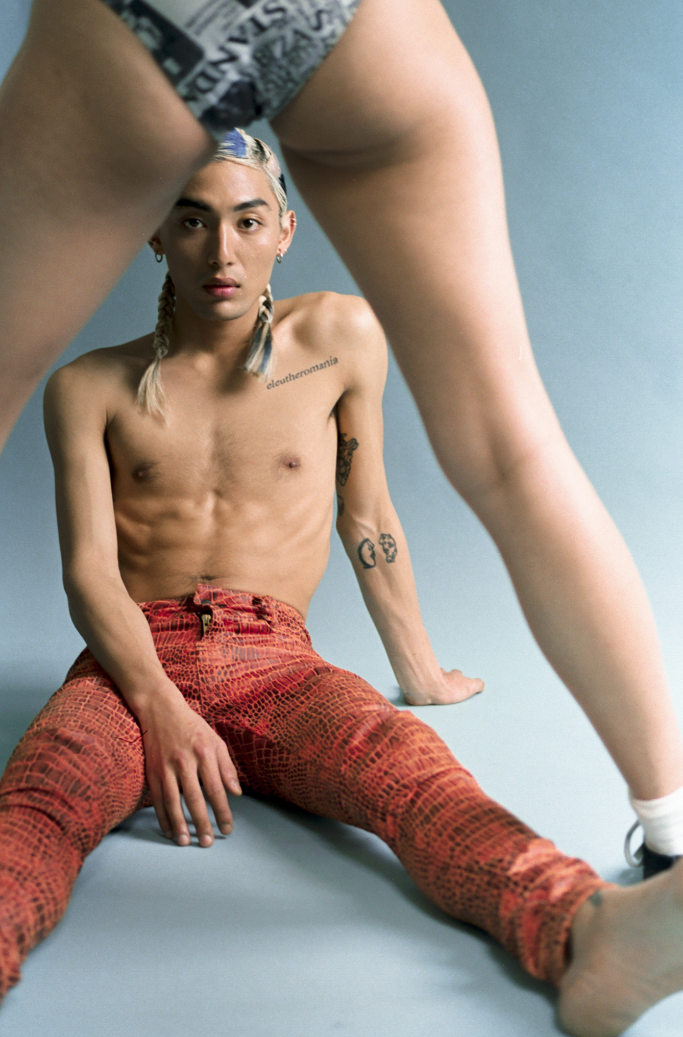 Shirtless Asian male poses with legs of a second human wearing newspaper-print shorts, set against a neutral backdrop.