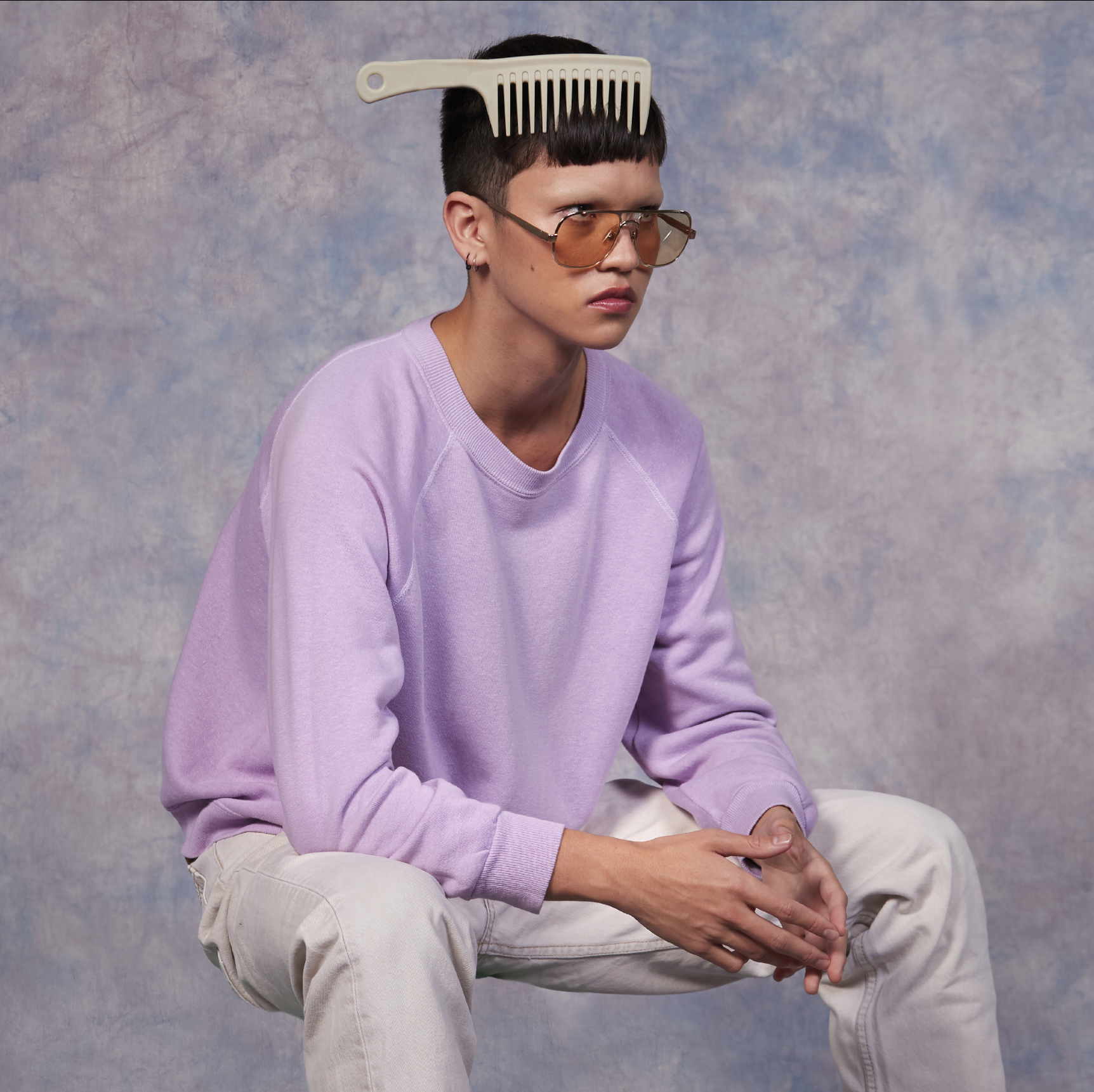 Person with comb on head poses indoors. Adult with short hair, glasses, and lavender sweatshirt sits against a mottled backdrop.