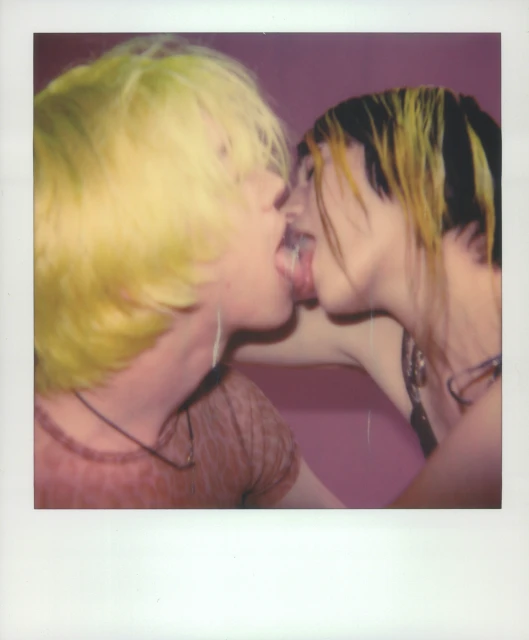 Close-up shot of two people with dyed hair kissing, tongues touching, against a solid pink background. Polaroids frame adds a vintage aesthetic.
