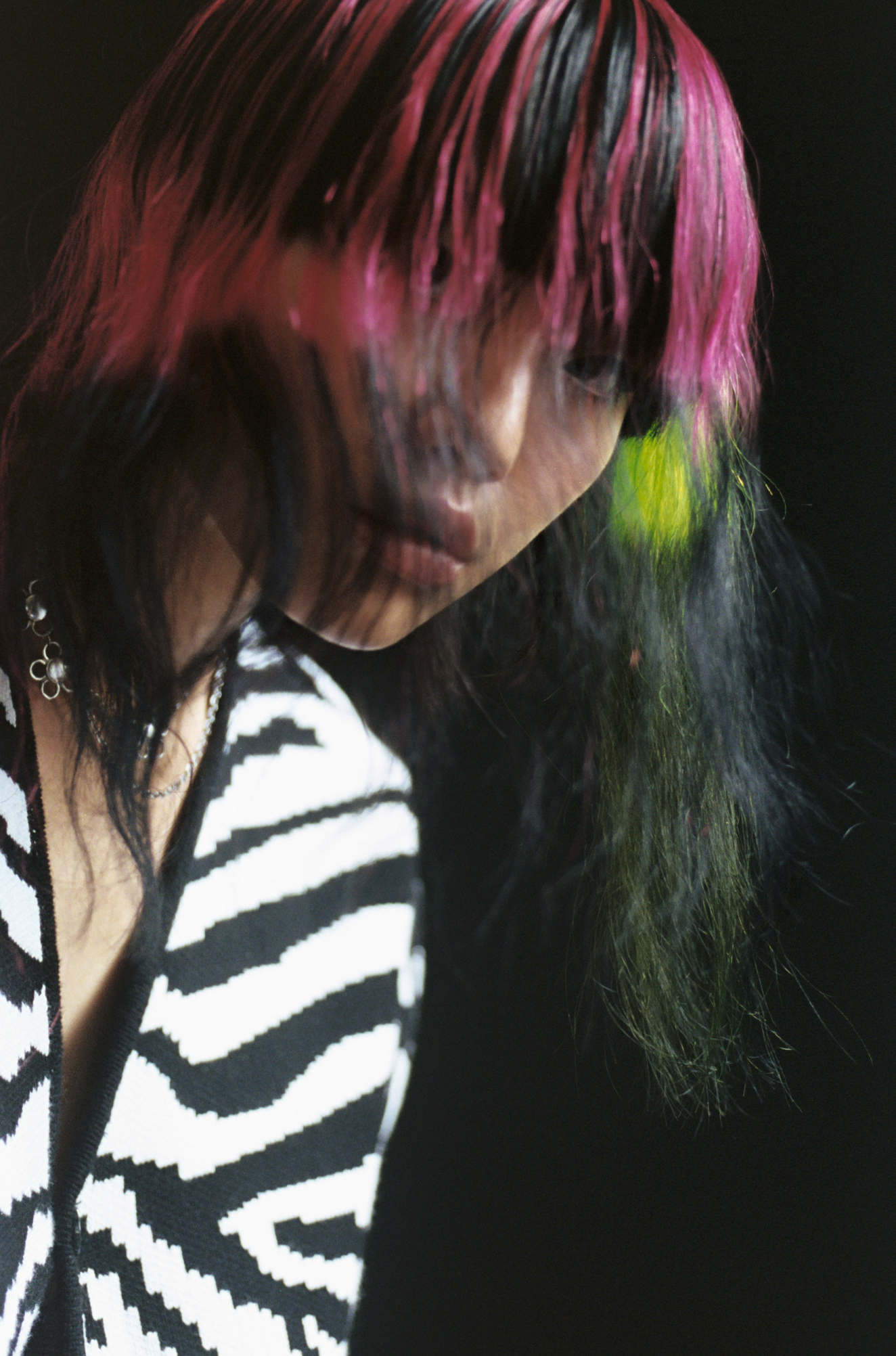 Portrait of an adult female with pink-streaked hair wearing a black and white patterned cardigan against a dark background.