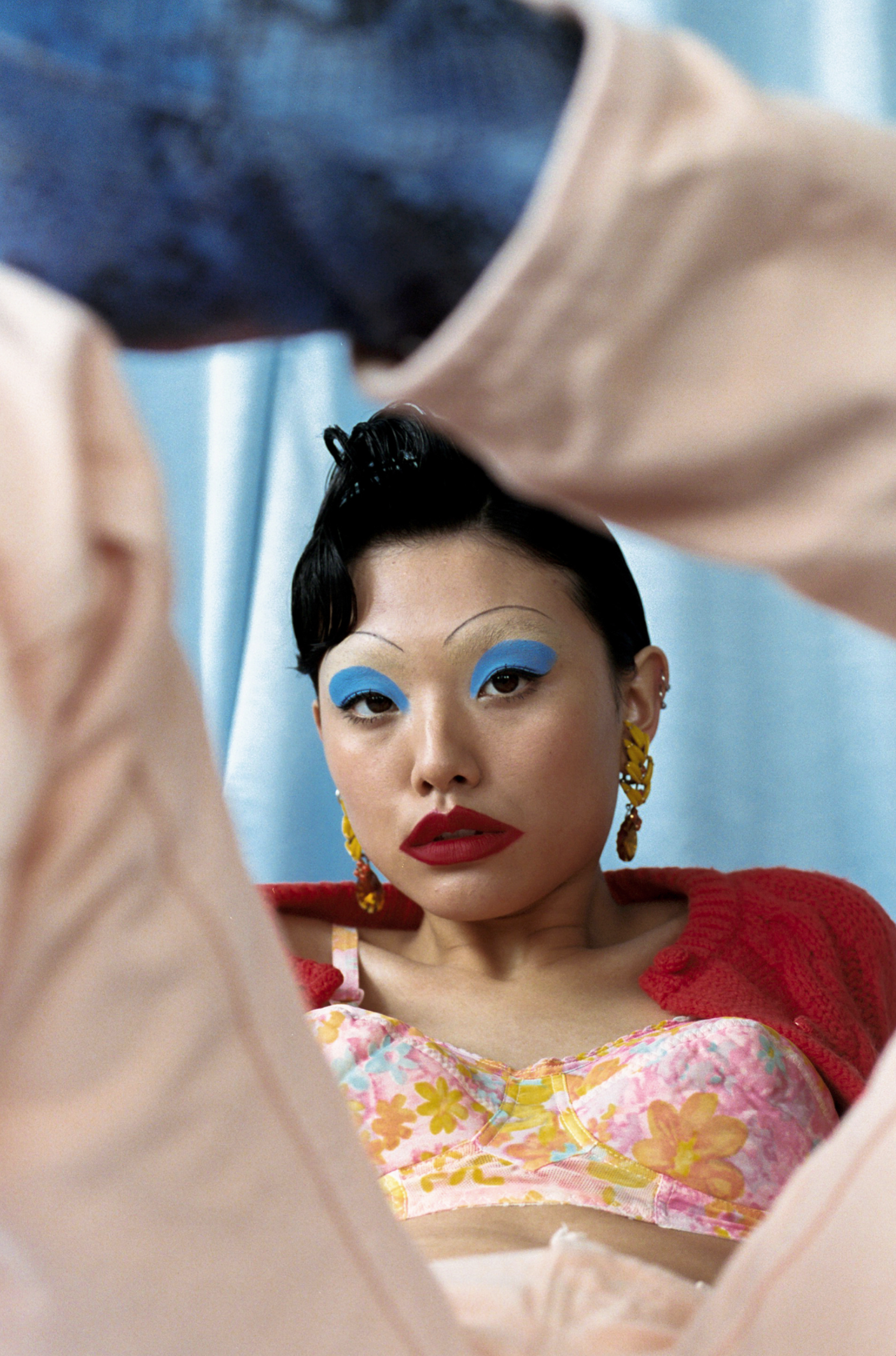 Portrait of an Asian woman with blue eyeshadow and bold red lipstick, wearing a floral bra top and red cardigan, studio shot.