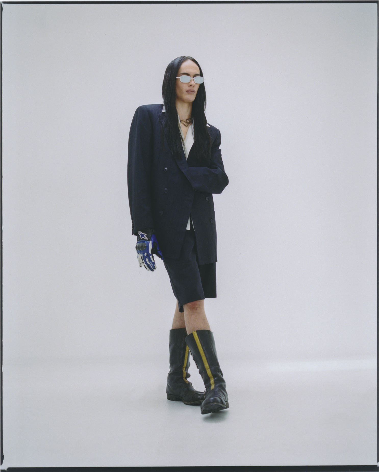 Woman with long dark hair poses in oversized navy blazer, matching shorts, futuristic sunglasses, and black and yellow striped boots in studio.