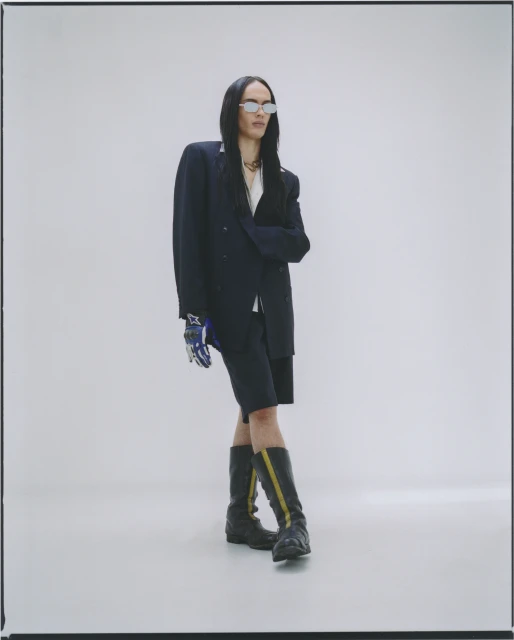 Woman with long dark hair poses in oversized navy blazer, matching shorts, futuristic sunglasses, and black and yellow striped boots in studio.