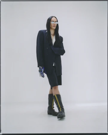 Woman with long dark hair poses in oversized navy blazer, matching shorts, futuristic sunglasses, and black and yellow striped boots in studio.
