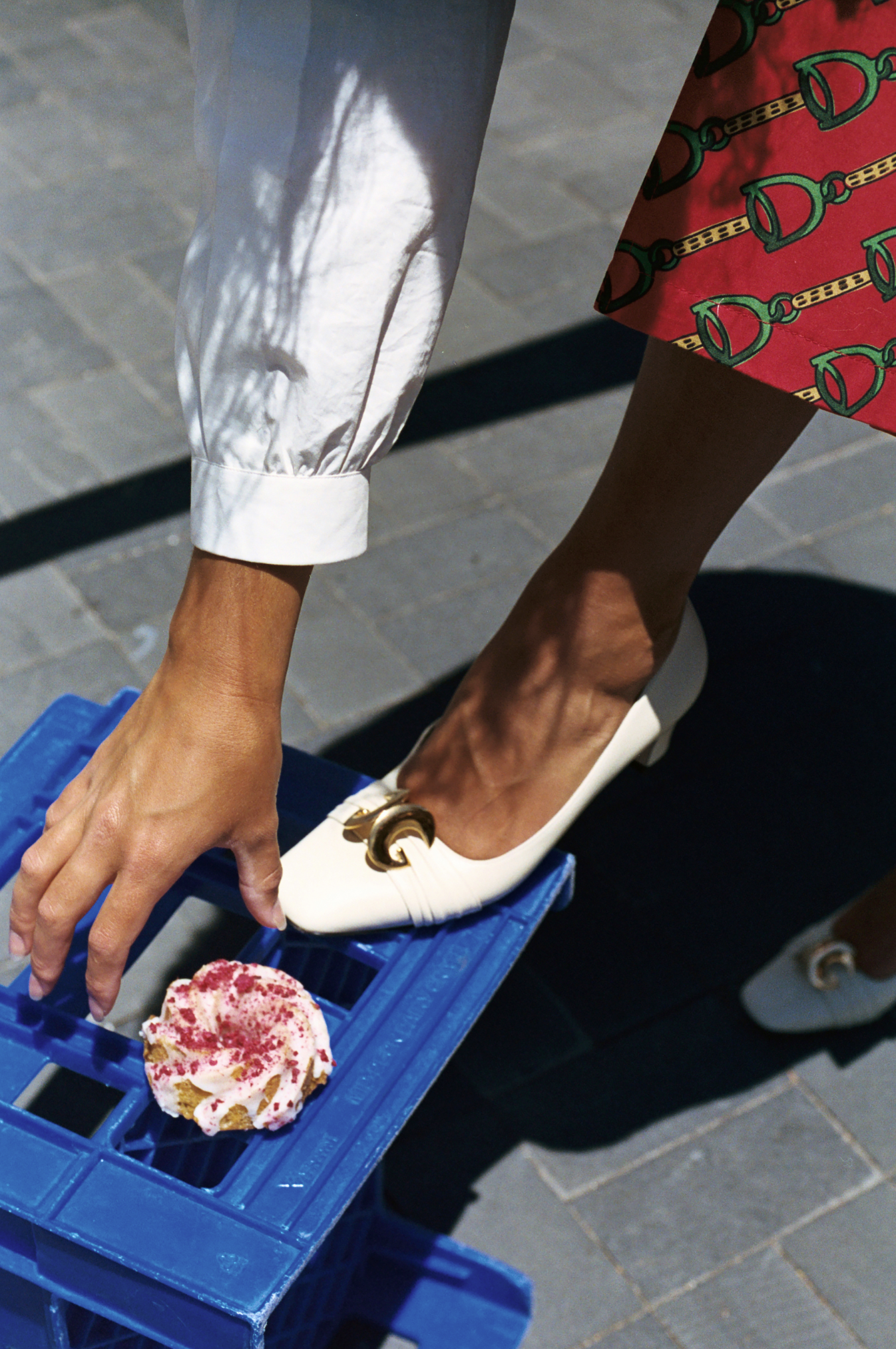 Person reaches for a frosted cupcake on a blue crate next to white shoes and a patterned red skirt, highlighting fashion and food.