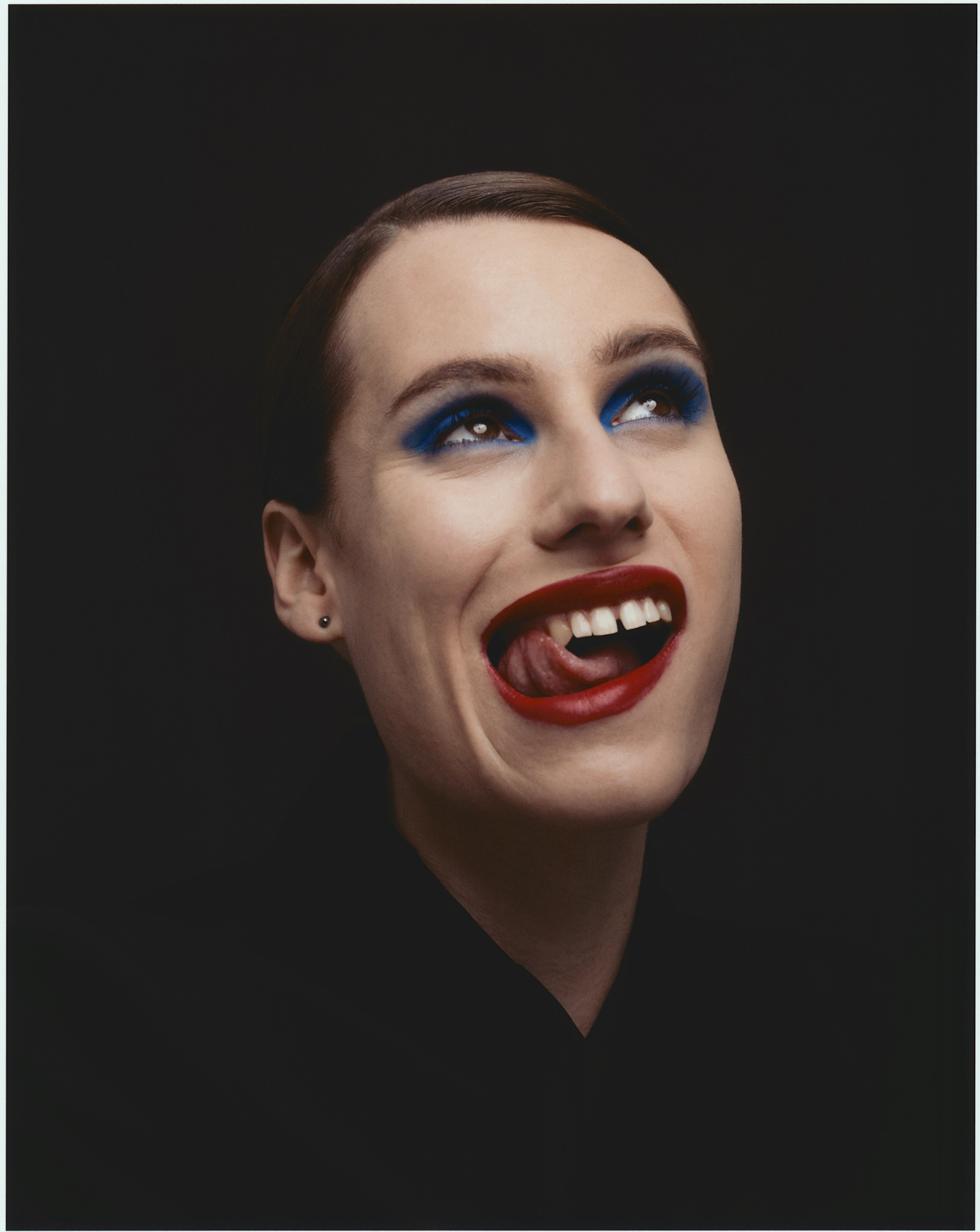 Close-up portrait of a smiling adult female with red lipstick and blue eyeshadow looking up against a black background.