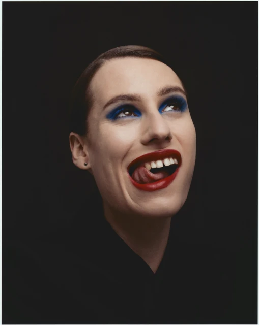 Close-up portrait of a smiling adult female with red lipstick and blue eyeshadow looking up against a black background.