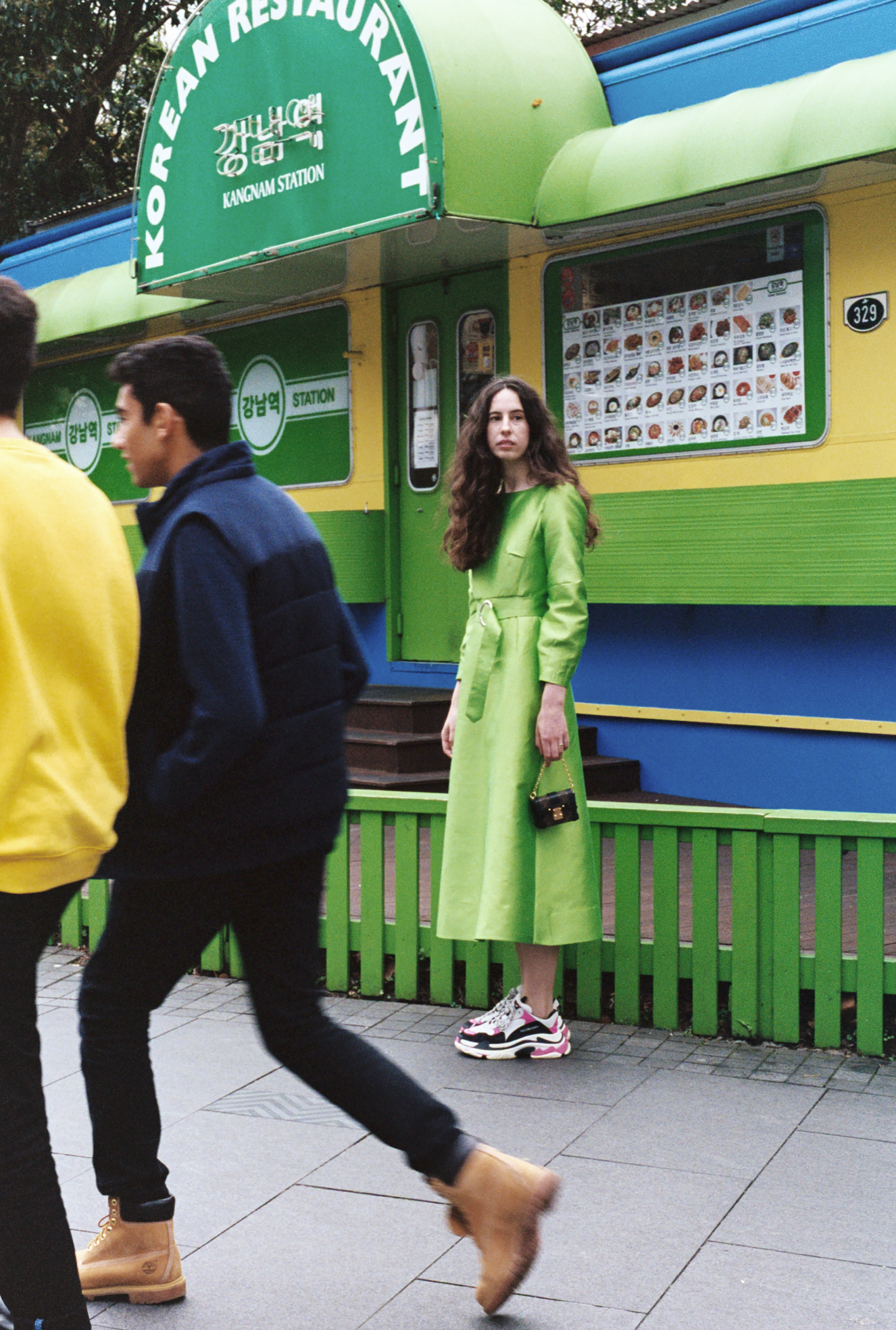 Woman in a green dress standing in front of a Korean restaurant as pedestrians walk on the sidewalk in an outdoor urban setting.