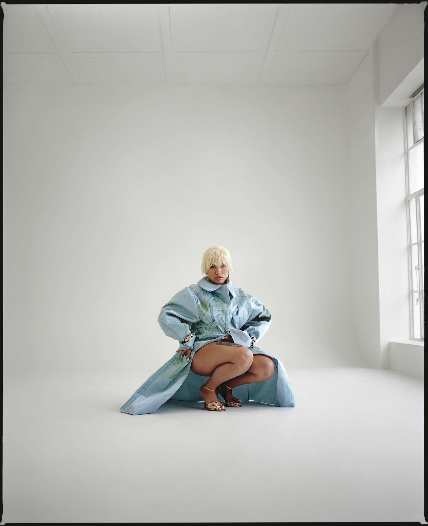 Full shot of a woman with blond hair crouching in a light blue coat and gold high-heeled sandals in a minimalist white studio setting.