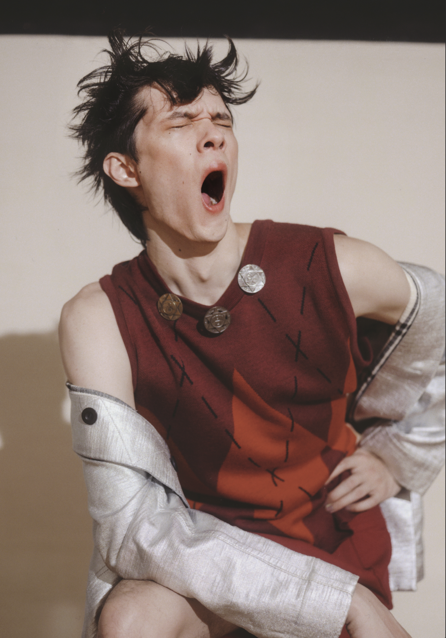 Portrait of a young adult with open mouth, wearing a burgundy sleeveless top and a silver jacket posing indoors.