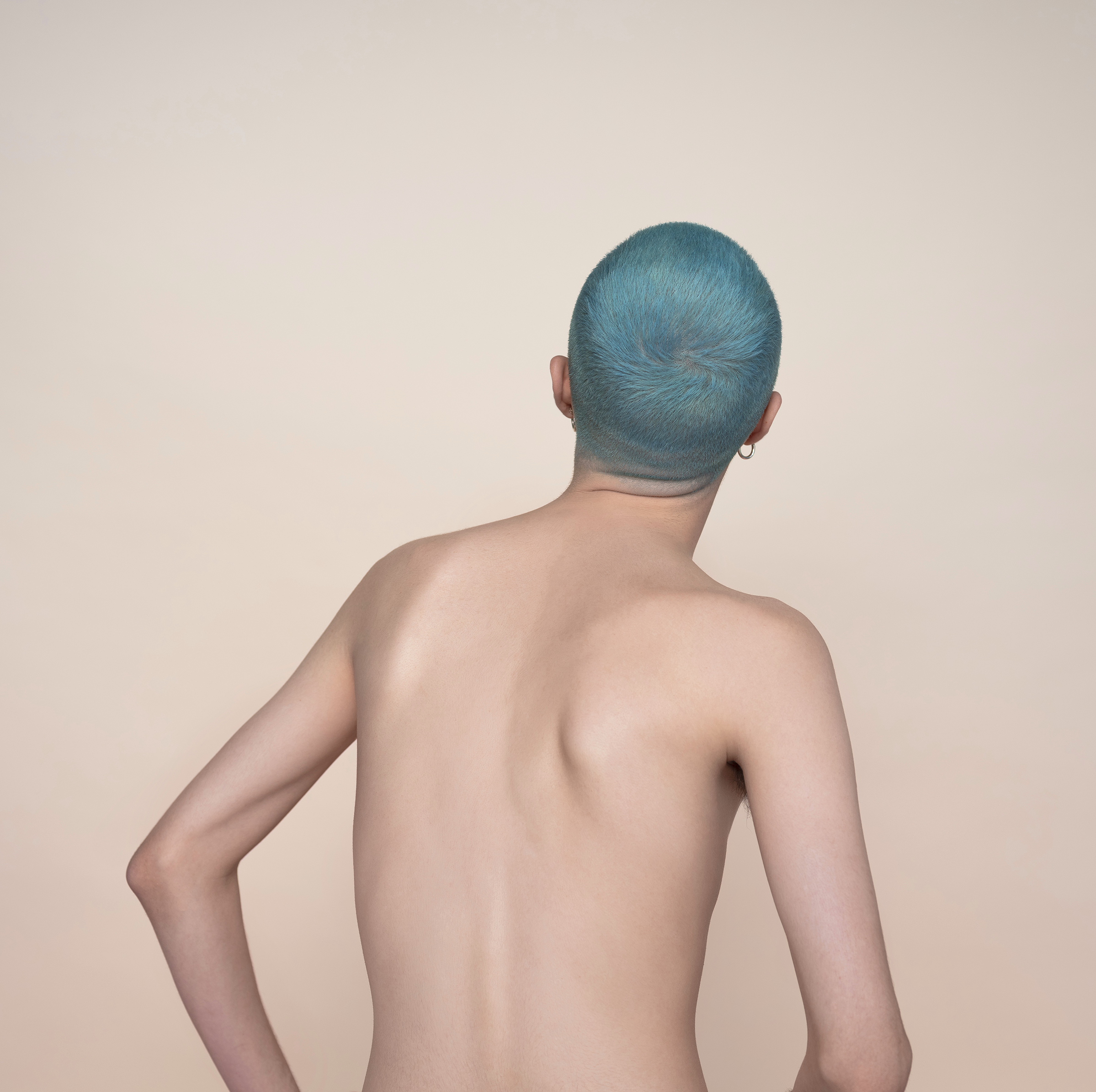 Close-up studio shot from behind of a slim adult with short, turquoise hair and fair skin against a tan backdrop. Minimalist.