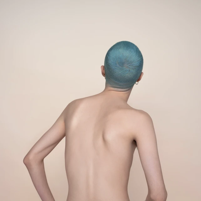 Close-up studio shot from behind of a slim adult with short, turquoise hair and fair skin against a tan backdrop. Minimalist.