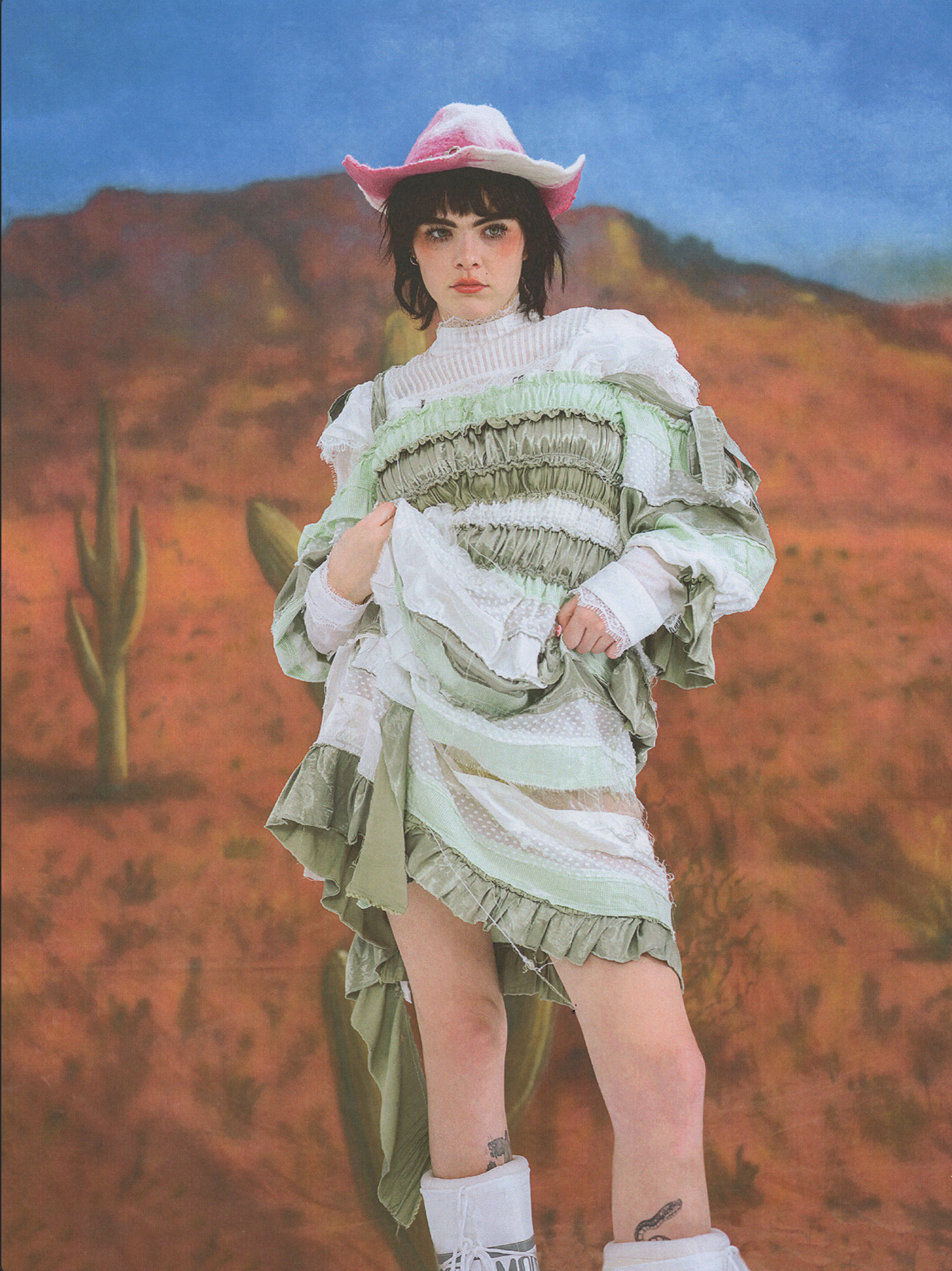 Brunette woman in a cowboy hat and green and white dress standing in front of a desert scene backdrop, posing for a fashion shot.