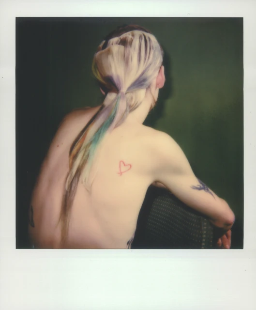 Back view of a light-skinned adult female with blonde and rainbow-colored hair tied back in a ponytail and a heart tattoo on her back.
