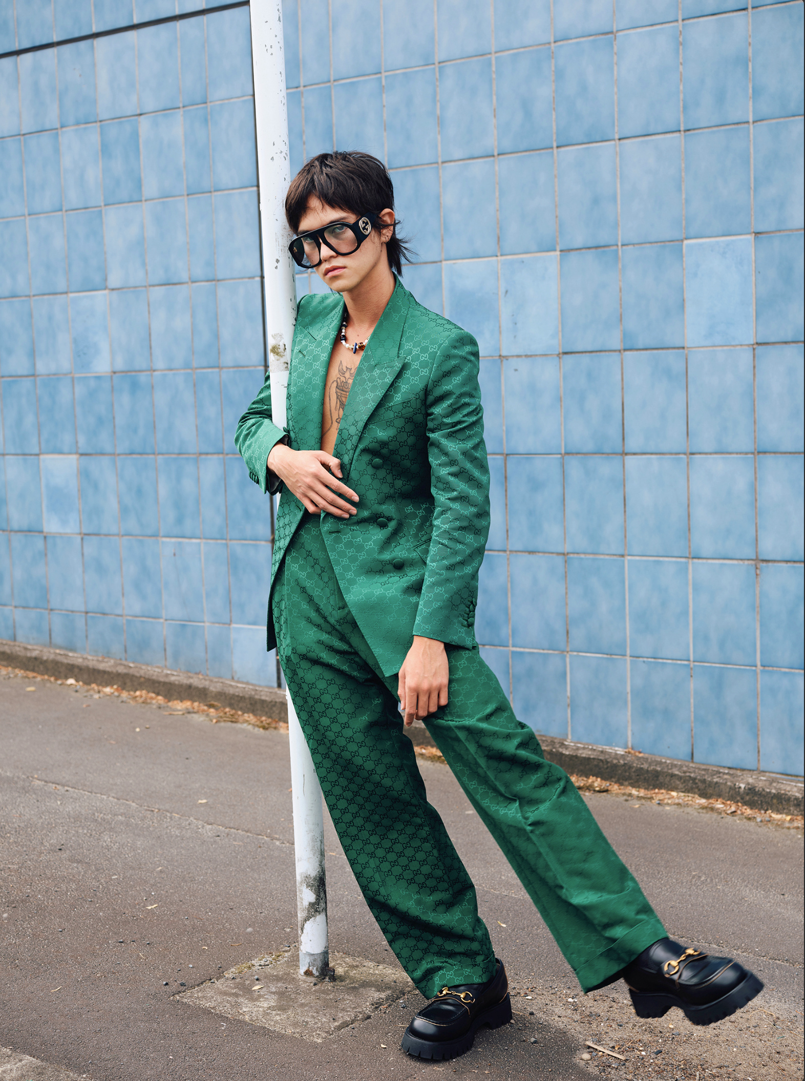A person poses in a green patterned suit, oversized glasses, and platform shoes against a blue tiled wall outdoors in an eye-level shot.
