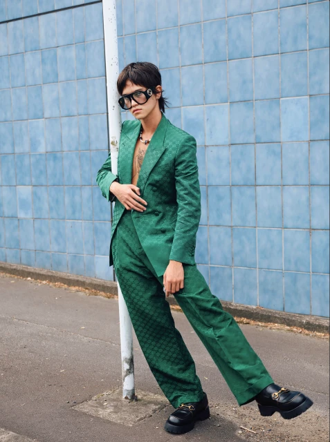 A person poses in a green patterned suit, oversized glasses, and platform shoes against a blue tiled wall outdoors in an eye-level shot.
