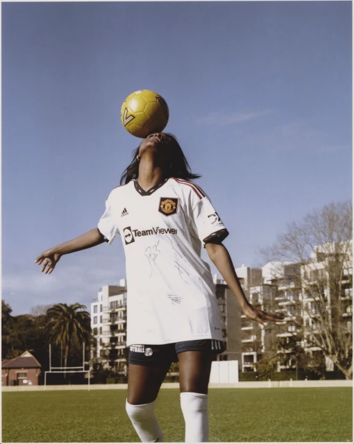 A woman balances a soccer ball on her head wearing a white Manchester United jersey on a green field under a blue sky.