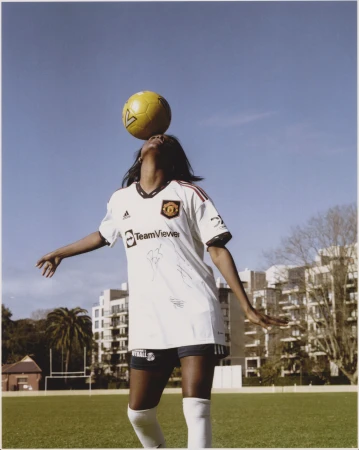 A woman balances a soccer ball on her head wearing a white Manchester United jersey on a green field under a blue sky.
