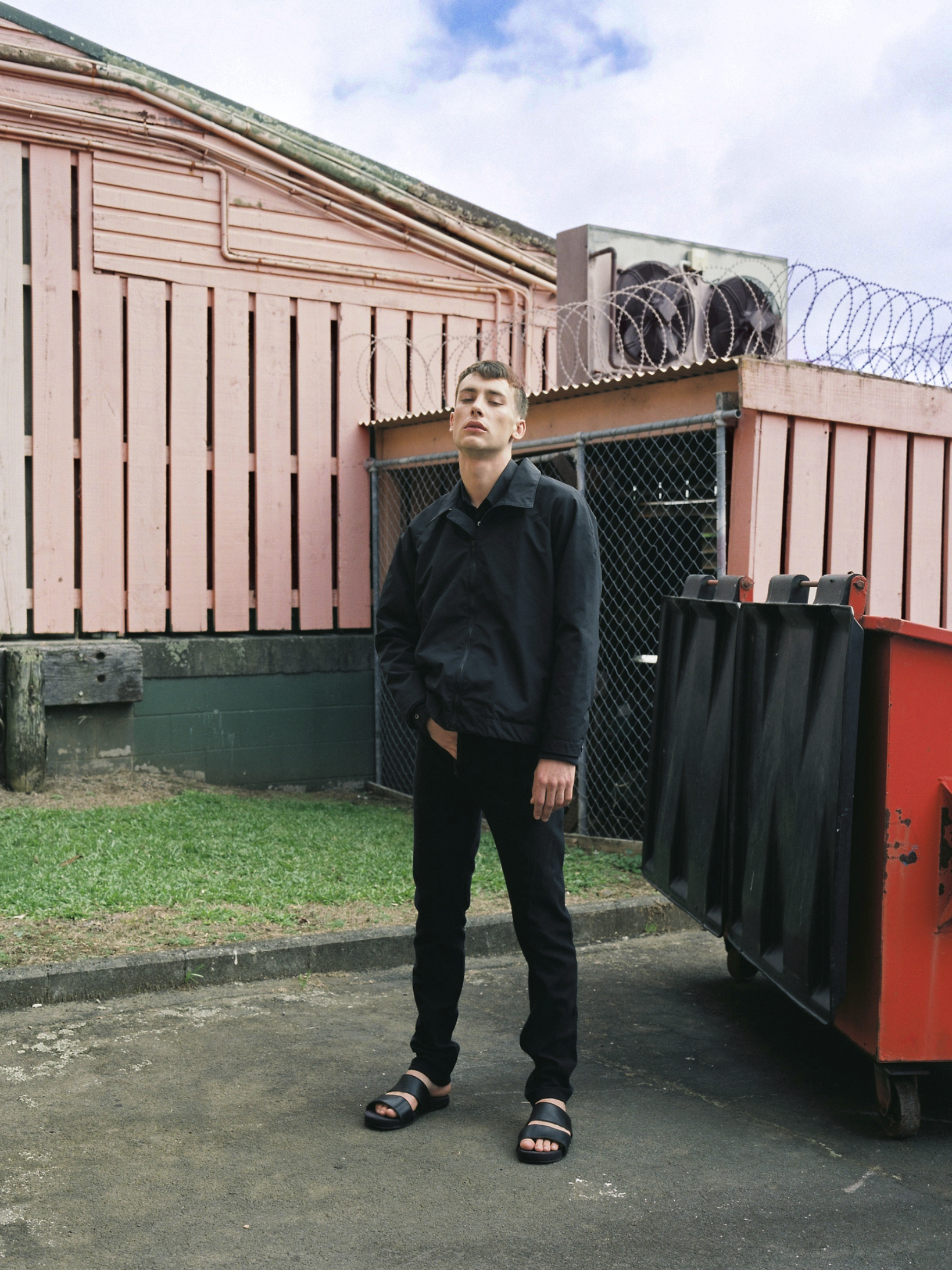 Young adult man in black jacket poses in industrial setting near a pink building and dumpster on concrete with a cloudy sky above.