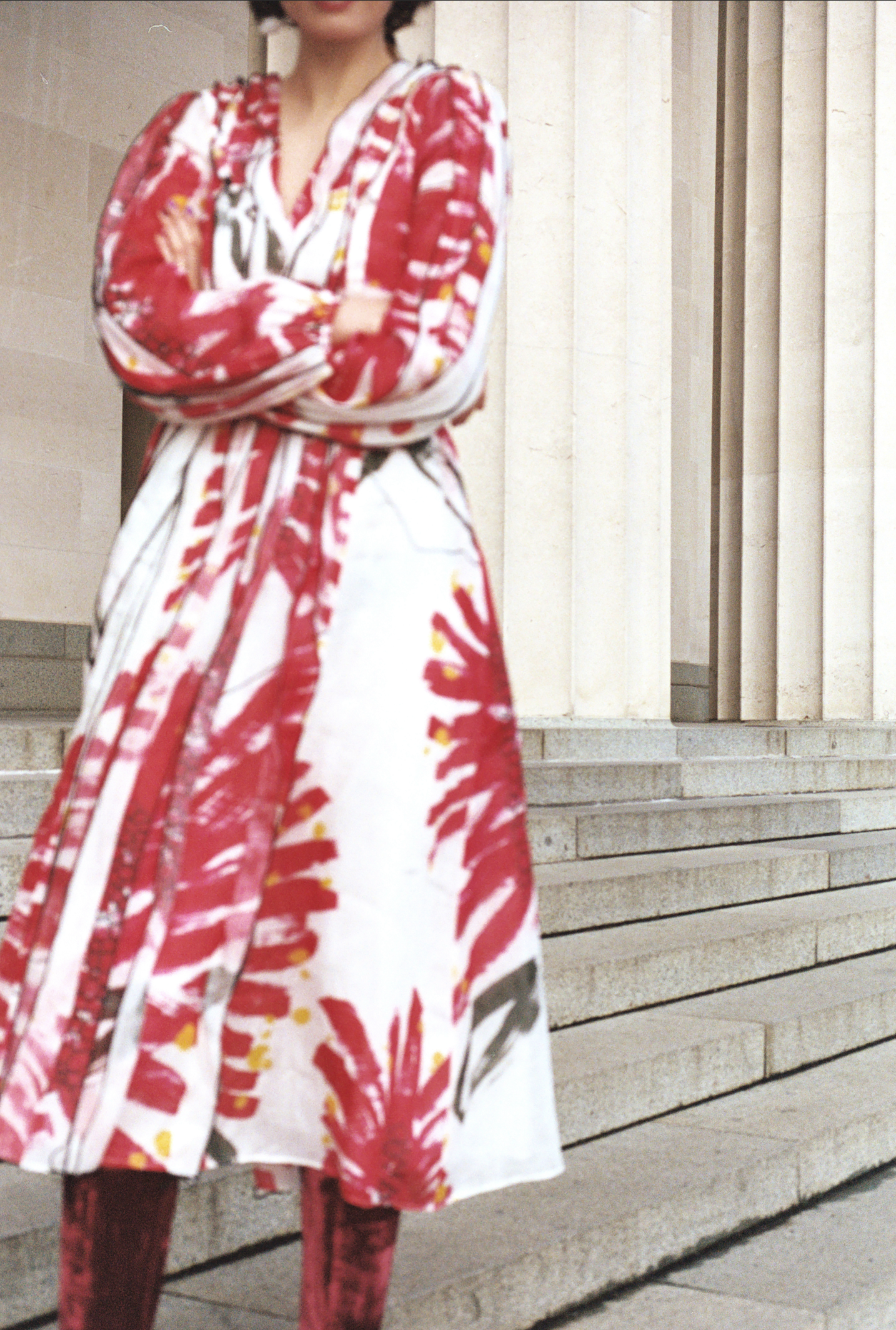 An adult female stands with crossed arms wearing a long white dress with an abstract red pattern. She is standing outside on stone steps.