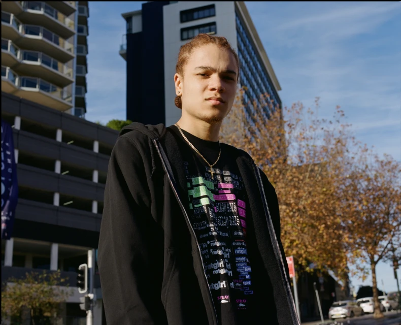 Portrait of a young adult man in black hoodie outdoors with city buildings, blue sky, and foliage on trees in the background.