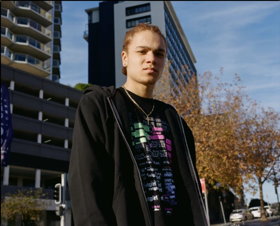 Portrait of a young adult man in black hoodie outdoors with city buildings, blue sky, and foliage on trees in the background.