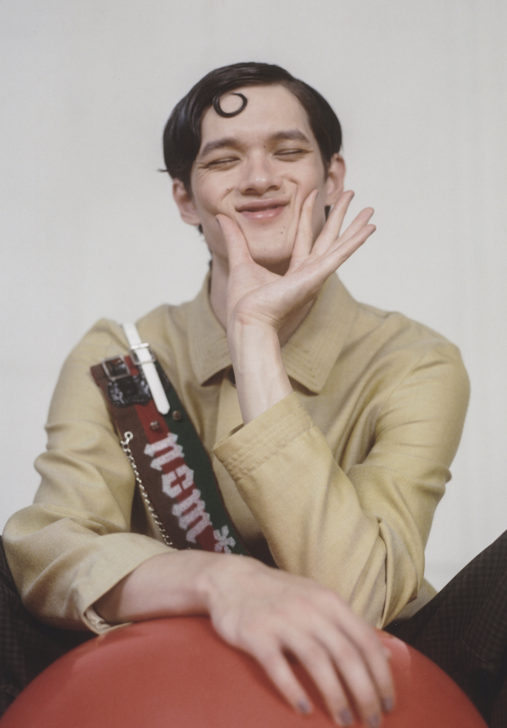 Portrait of an adult male with a decorative handbag, smiling slightly and holding his hand to his face, in a studio setting.