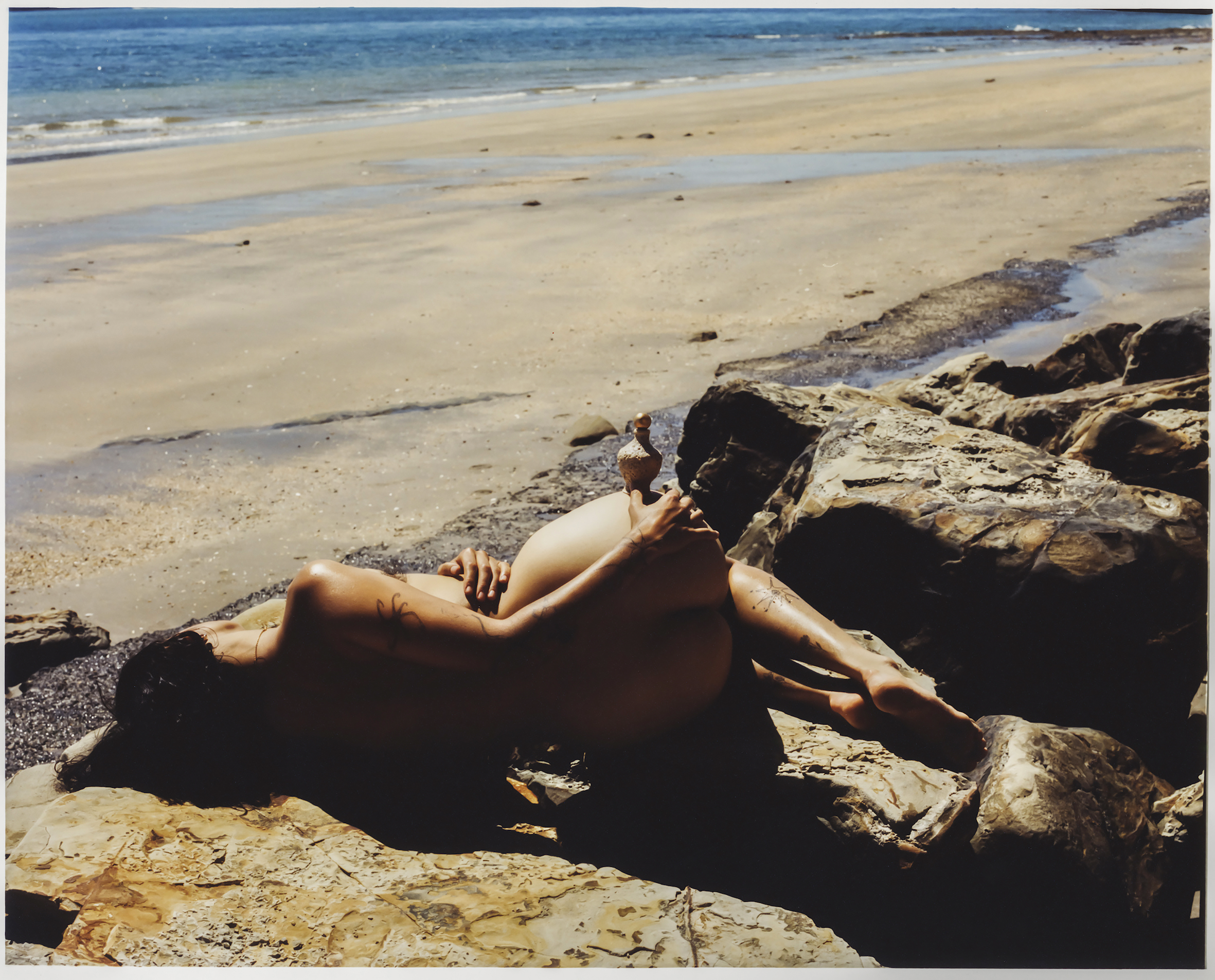 Nude adult female rests on rocks by the beach, holding a vase. The scene captures the beach shoreline and ocean in the background.