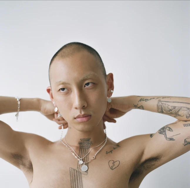 Portrait of a bald, androgynous person with visible tattoos and silver jewelry posing indoors against a bright, plain background.