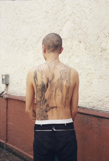 Man with a shaved head and tattoos on his back and head stands with his back to the camera in front of a textured wall.