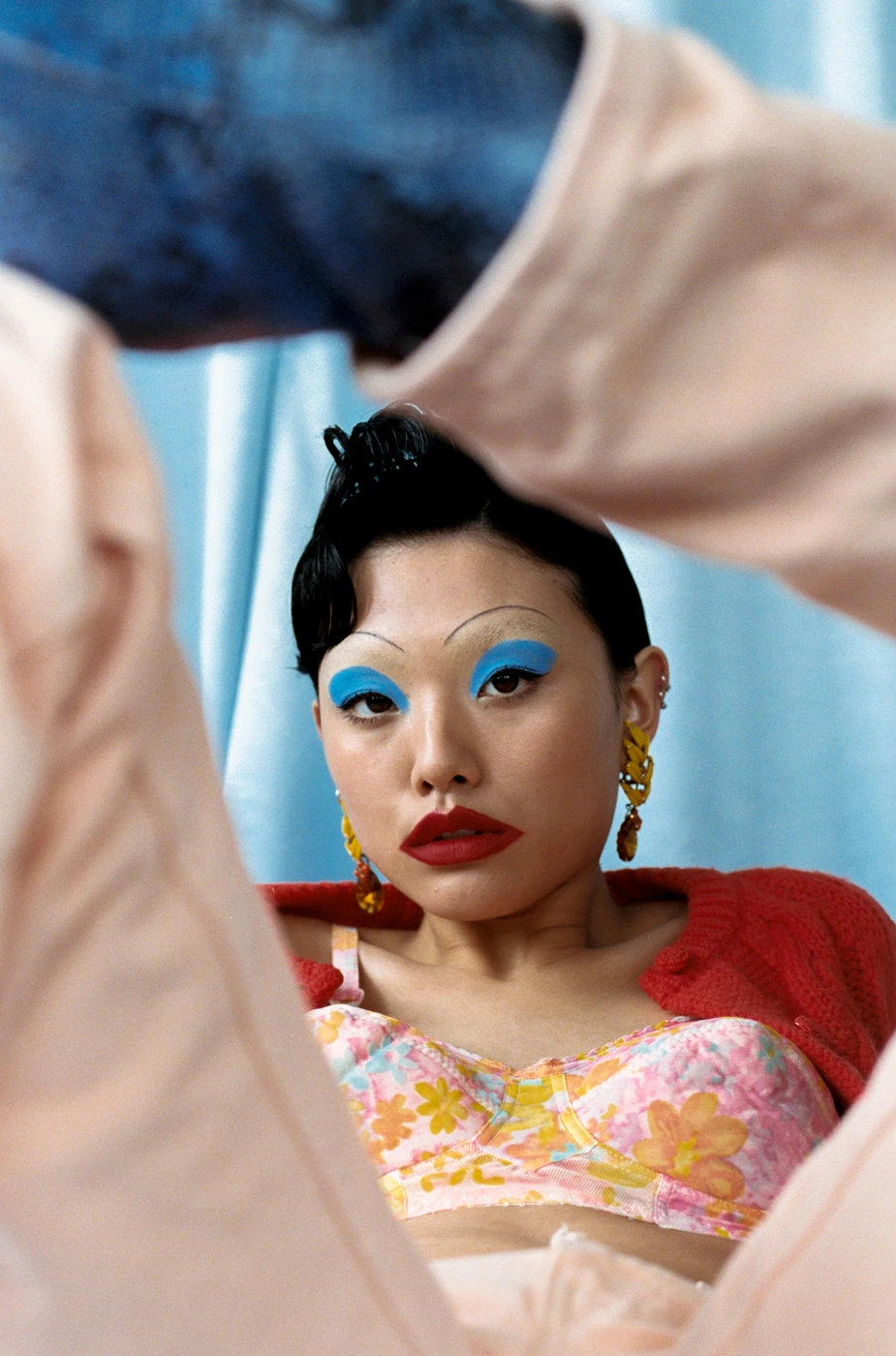 Portrait of a woman with bold blue eyeshadow, red lipstick, and thin arched eyebrows framed by limbs in the foreground.