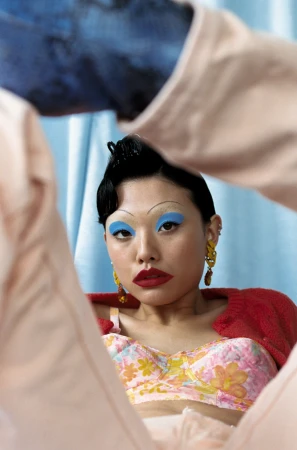 Portrait of a woman with bold blue eyeshadow, red lipstick, and thin arched eyebrows framed by limbs in the foreground.