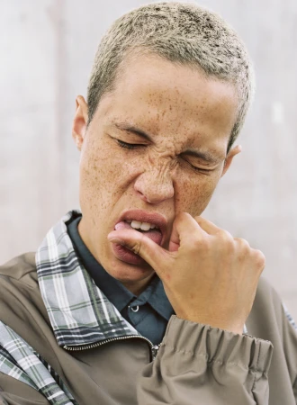 Close-up portrait of a person with short hair and freckles pulling down their lower lip against a neutral background.