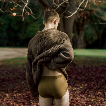 A person viewed from behind in an outdoor wooded area wearing a chunky brown knit sweater pulled up and olive green briefs