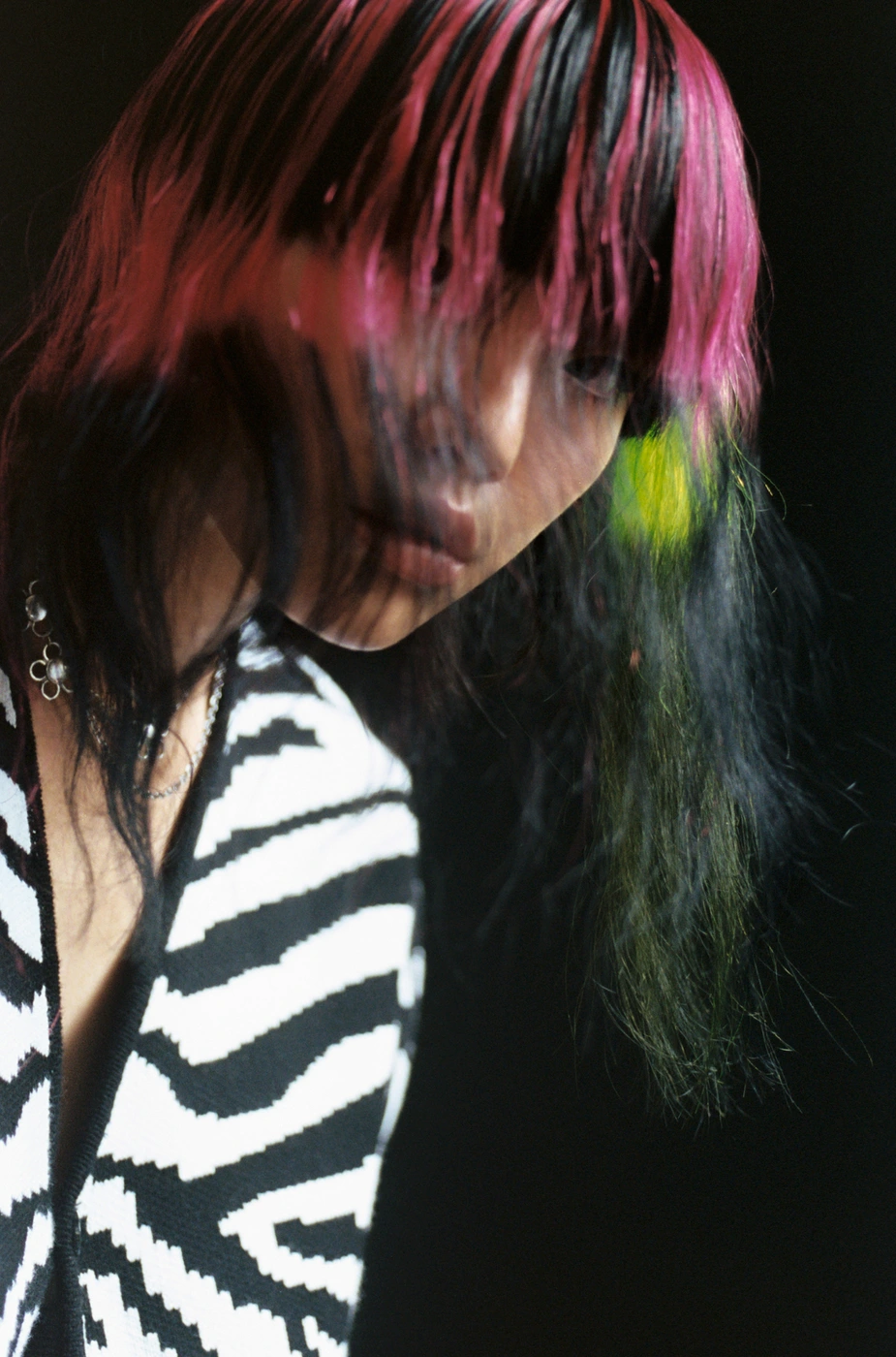 Portrait of a person with dyed pink and black hair wearing a black and white zebra print top against a dark background.