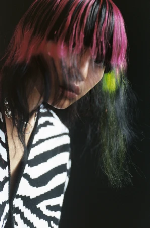 Portrait of a person with dyed pink and black hair wearing a black and white zebra print top against a dark background.