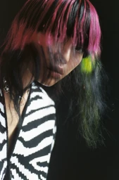Portrait of a person with dyed pink and black hair wearing a black and white zebra print top against a dark background.