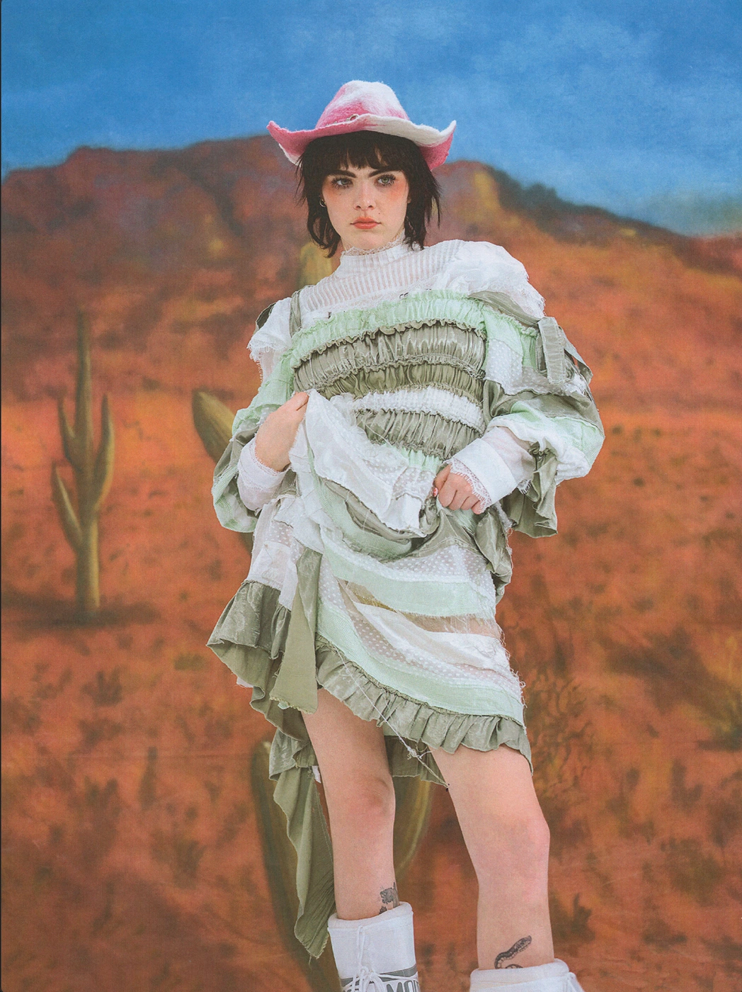 Model wearing a ruffled sage green dress and pink cowboy hat against a painted desert landscape backdrop.