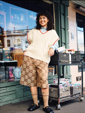 Smiling woman wearing a cream cable-knit vest, patterned shorts, and black loafers leaning against a book cart outside.
