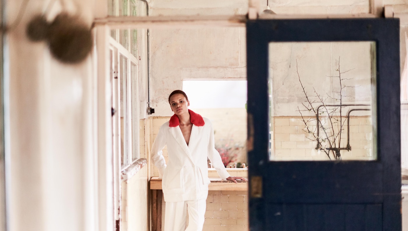 Woman in a white corduroy suit with a red collar poses in a distressed interior with a partially visible door and window frames.