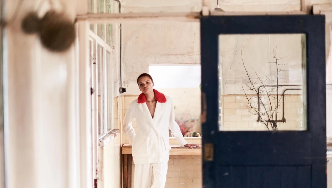 Woman in a white corduroy suit with a red collar poses in a distressed interior with a partially visible door and window frames.