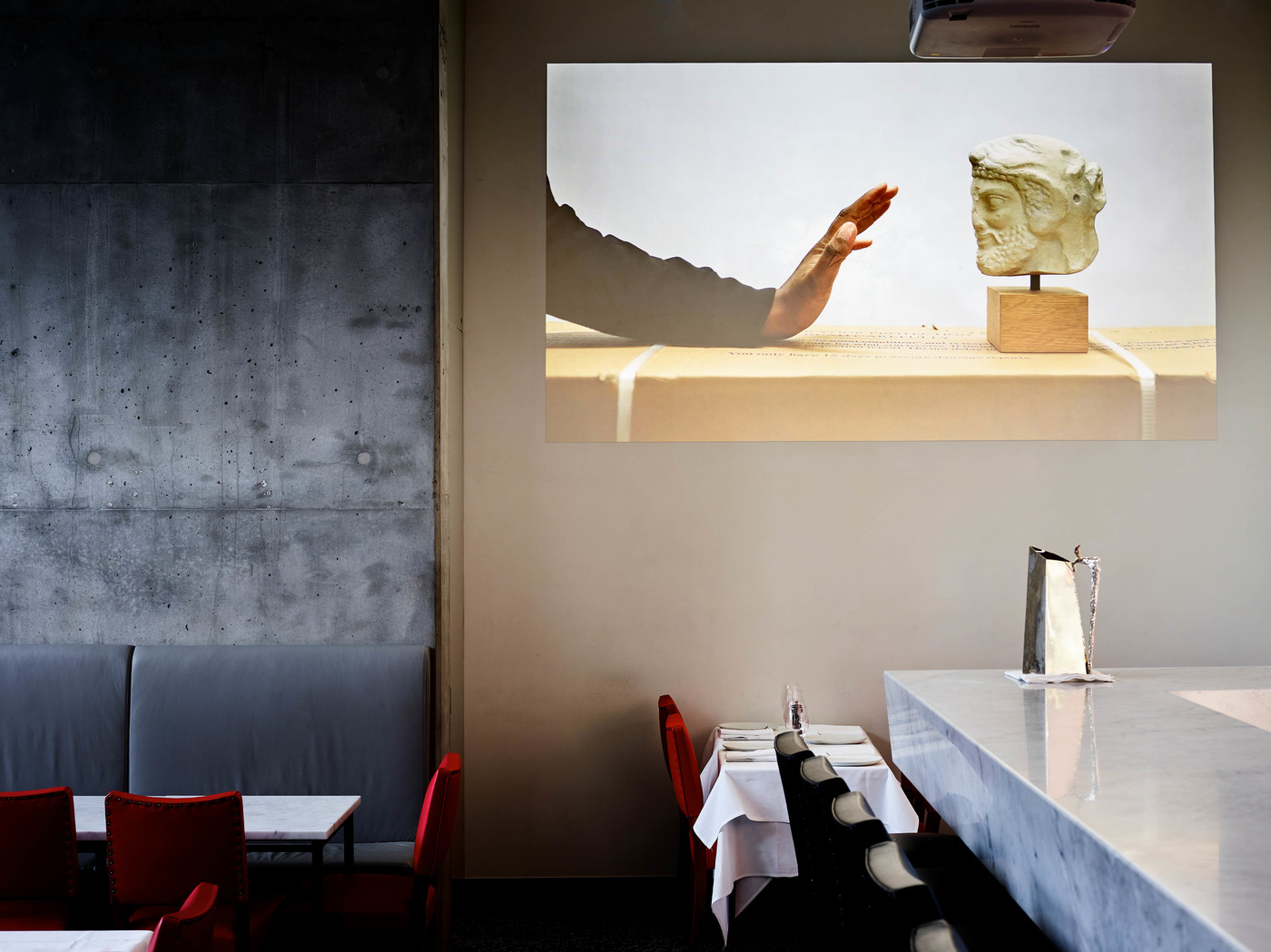 Artwork in a restaurant featuring a photograph of a hand reaching towards a Greek sculpture bust on a beige surface against a white backdrop.