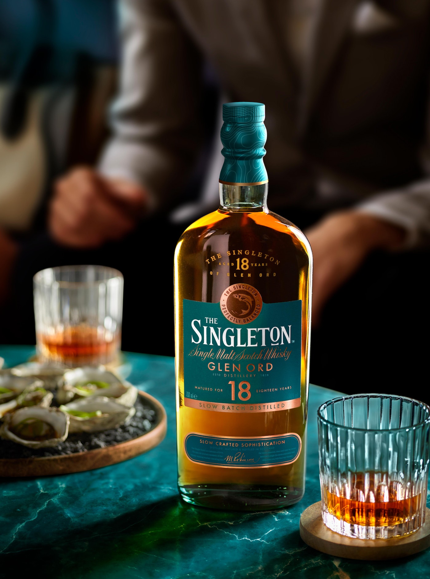 Singleton 18-year Single Malt Scotch Whisky bottle with two glasses, oysters, and an out-of-focus person in the background.