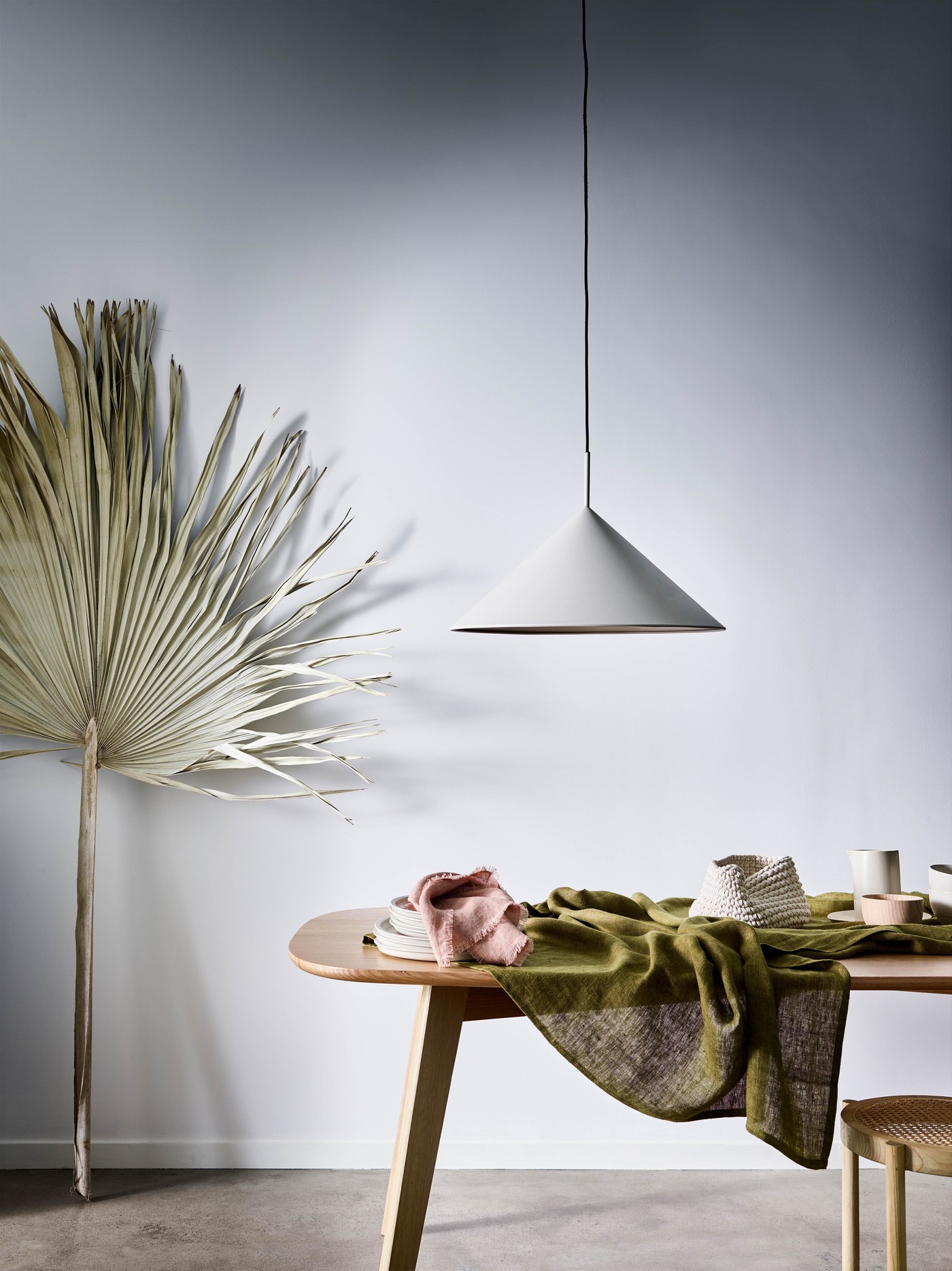 Interior scene featuring a cone pendant light above a dining table with linens, a palm frond, and a cane chair against a white wall.