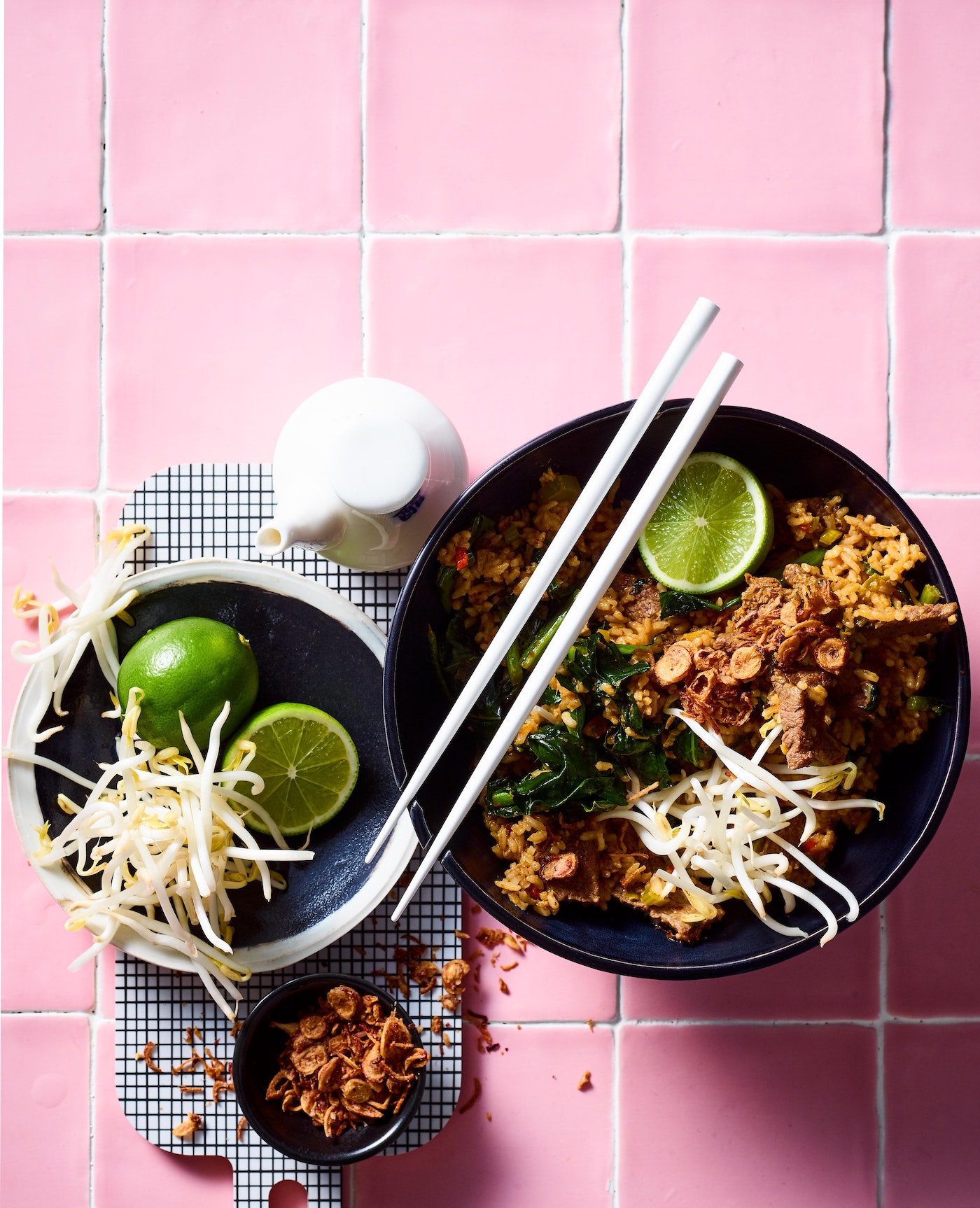 Overhead shot of a vibrant Asian-inspired noodle bowl garnished with fresh lime, bean sprouts, and crispy toppings on a pink tiled surface.