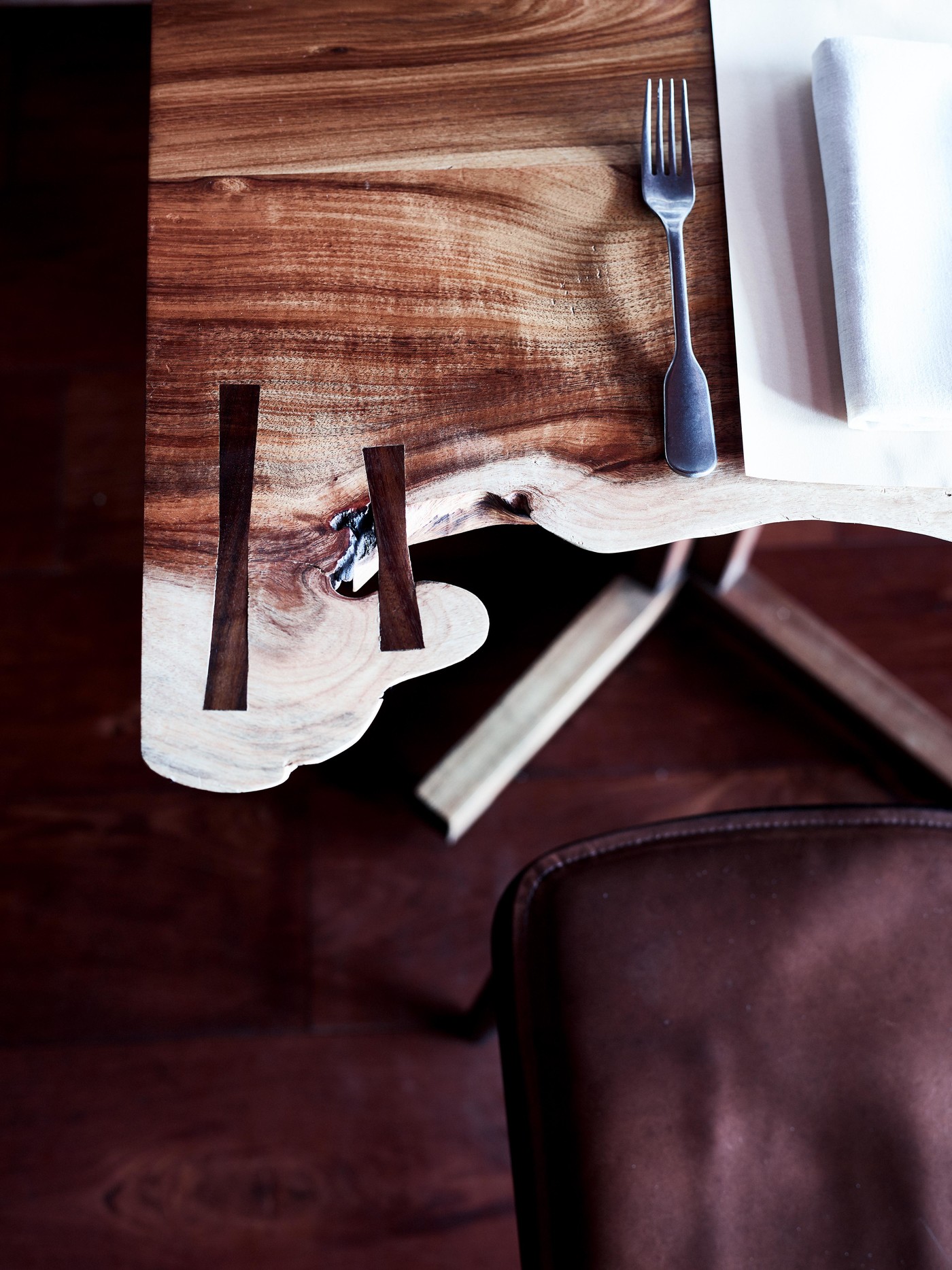 Close-up of a place setting on a live edge wood table, including a fork, linen napkin, and chair. Brown wooden floor in the background.