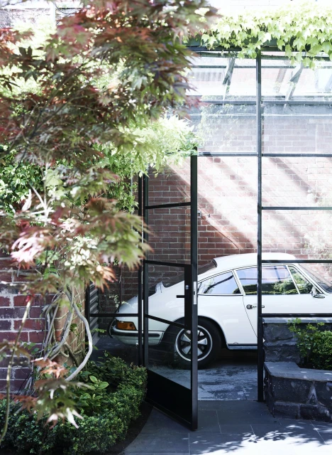 Outdoor view of a white Porsche through a black-framed glass door, with greenery and a brick wall adding to the aesthetic.